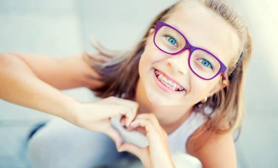 Smiling little girl with braces and glasses showing heart with hands